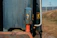 A close-up of an industrial object with a prominent blue casing featuring a yellow lightning bolt symbol. A red cylindrical part is visible below the blue component. The background includes a metallic surface and a distant view of a grassy area with power lines.