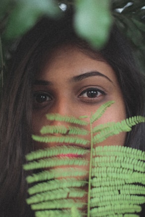 A woman's face is partially obscured by a green fern leaf, with an emphasis on her eyes and brow. The composition creates an intimate and natural feel, with rich, earthy tones.