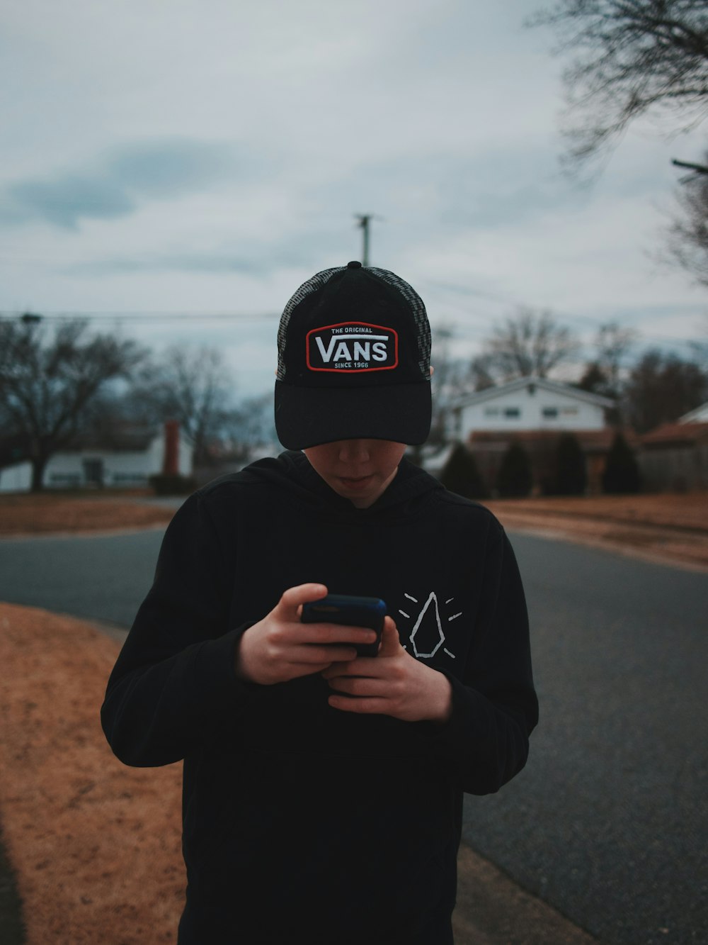 person in black Vans cap and black sweater holding smartphone white standing beside road
