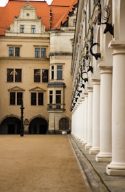 An architectural scene featuring a stately building with multiple stories and an orange-red tiled roof. The facade displays a series of windows and decorative stonework. The right side of the image presents a line of white columns supporting an arcade, with mounted busts of animals above them. The ground is paved with sand or dirt, and an ornate black lamp post is visible.