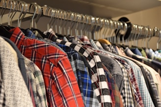 A row of plaid and patterned shirts on hangers, closely arranged and displayed on a clothing rack. The fabrics vary in color and design, featuring a mix of reds, blues, and black checks.
