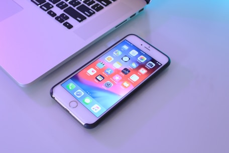 A smartphone with a colorful home screen lies on a flat surface near the edge of a laptop. The phone's screen displays various app icons organized in a grid pattern. The laptop is partially open, showing the keyboard and part of the screen. Soft lighting reflects off the surfaces, creating a calming ambiance.