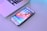 A smartphone with a colorful home screen lies on a flat surface near the edge of a laptop. The phone's screen displays various app icons organized in a grid pattern. The laptop is partially open, showing the keyboard and part of the screen. Soft lighting reflects off the surfaces, creating a calming ambiance.