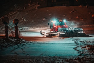 A snow plow is actively working on a snow-covered road at night, illuminated by headlights. The surroundings include snowbanks and a couple of signs on the left side. The scene is dimly lit with an atmospheric glow from the plow's lights.