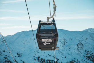 A gondola lift is suspended in the air against a backdrop of snow-covered mountains. The ski lift cabin, marked with branding for Meribel, is part of a cable car system set in a clear blue sky with a scattering of clouds. The scene captures the serene and cold environment of a mountainous ski resort.