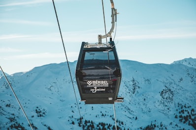 A gondola lift is suspended in the air against a backdrop of snow-covered mountains. The ski lift cabin, marked with branding for Meribel, is part of a cable car system set in a clear blue sky with a scattering of clouds. The scene captures the serene and cold environment of a mountainous ski resort.