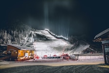 A nighttime winter sports scene with a well-lit ski slope, lined with trees on either side, attracting people at the base. The artificial lighting illuminates the snow-covered landscape, creating a vibrant atmosphere. Spectators and possible participants gather near a building on the left, possibly a ski lodge or rental station.