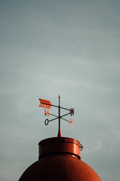 A metal weather vane sits atop a rounded structure, silhouetted against a cloudy sky. The weather vane features an arrow and directional letters (N, E, S, W) to indicate wind direction.