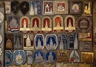 A detailed collection of vintage pins and badges displayed on a board. Each piece features intricate designs and inscriptions, many of which depict buildings and towers. The colors used are primarily gold, red, blue, and silver, giving the collection a historical and regal appearance.