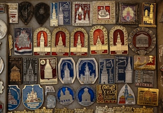 A detailed collection of vintage pins and badges displayed on a board. Each piece features intricate designs and inscriptions, many of which depict buildings and towers. The colors used are primarily gold, red, blue, and silver, giving the collection a historical and regal appearance.