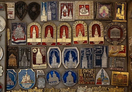 A detailed collection of vintage pins and badges displayed on a board. Each piece features intricate designs and inscriptions, many of which depict buildings and towers. The colors used are primarily gold, red, blue, and silver, giving the collection a historical and regal appearance.