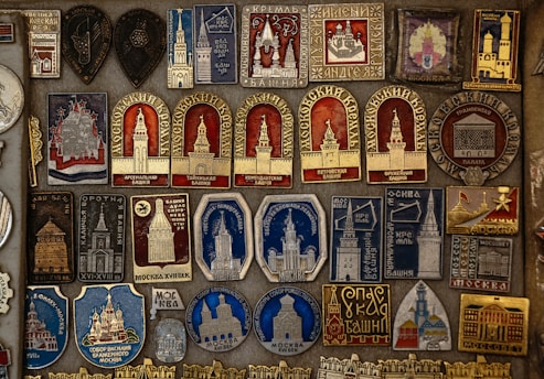 A detailed collection of vintage pins and badges displayed on a board. Each piece features intricate designs and inscriptions, many of which depict buildings and towers. The colors used are primarily gold, red, blue, and silver, giving the collection a historical and regal appearance.