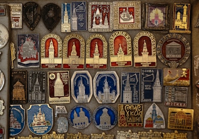 A detailed collection of vintage pins and badges displayed on a board. Each piece features intricate designs and inscriptions, many of which depict buildings and towers. The colors used are primarily gold, red, blue, and silver, giving the collection a historical and regal appearance.