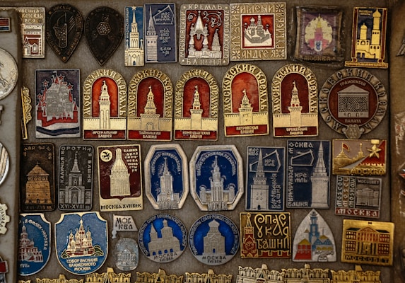 A detailed collection of vintage pins and badges displayed on a board. Each piece features intricate designs and inscriptions, many of which depict buildings and towers. The colors used are primarily gold, red, blue, and silver, giving the collection a historical and regal appearance.