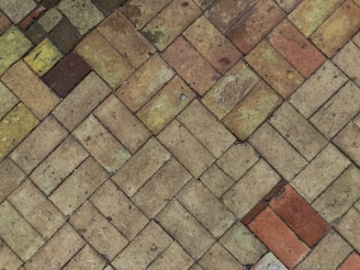 Interlocking bricks of varying earth tones form a herringbone pattern. The bricks display a range of hues from light tan to rusty red, with patches of moss and weathering visible. The texture appears rough and aged.