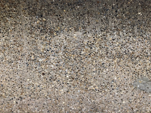 A close-up view of an aggregate concrete surface composed of a mix of small pebbles embedded in cement, creating a textured and rough appearance.