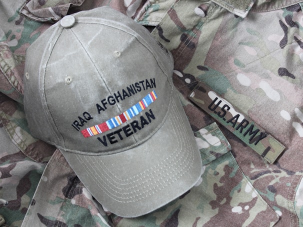 A khaki cap with the words 'Iraq Afghanistan Veteran' embroidered on it is placed on a camouflage military uniform that has a 'U.S. Army' tag.