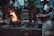 A person is forging metal with a small forge, where sparks and flames are visible. Several rods are being heated and manipulated by the person, while a group of people stands in the background observing. The scene appears to be outdoors.
