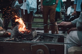A person is forging metal with a small forge, where sparks and flames are visible. Several rods are being heated and manipulated by the person, while a group of people stands in the background observing. The scene appears to be outdoors.