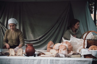 Two women are at a rustic market stall displaying various types of bread. One woman slices a loaf while wearing a headscarf, and the other woman sits looking away. The setting includes a clay jug and a large basket on a white tablecloth, with a backdrop of a plain, dark curtain.