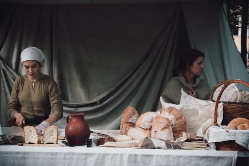 Two women are at a rustic market stall displaying various types of bread. One woman slices a loaf while wearing a headscarf, and the other woman sits looking away. The setting includes a clay jug and a large basket on a white tablecloth, with a backdrop of a plain, dark curtain.