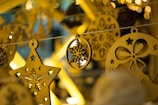 A close-up of intricately designed Christmas ornaments made of wood, featuring a star with cut-out snowflakes and an oval with a snowflake pattern, all hanging on a string. The background is filled with blurred, warm yellow lights creating a cozy and festive atmosphere.