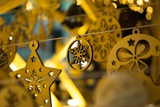 A close-up of intricately designed Christmas ornaments made of wood, featuring a star with cut-out snowflakes and an oval with a snowflake pattern, all hanging on a string. The background is filled with blurred, warm yellow lights creating a cozy and festive atmosphere.