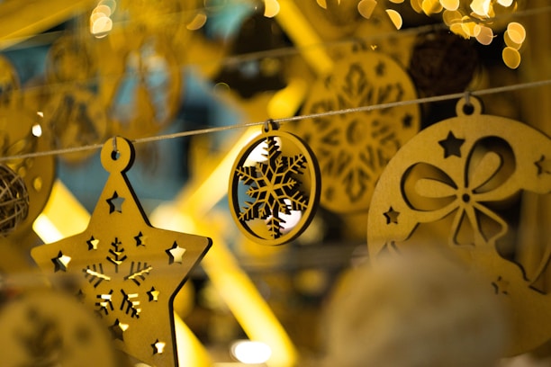 A close-up of intricately designed Christmas ornaments made of wood, featuring a star with cut-out snowflakes and an oval with a snowflake pattern, all hanging on a string. The background is filled with blurred, warm yellow lights creating a cozy and festive atmosphere.