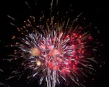 A spectacular fireworks display bursting in reds and golds above a concert stage with dynamic lighting and an excited crowd.