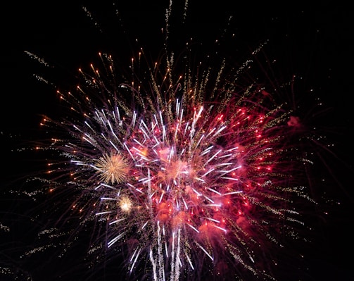 A vibrant display of fireworks with bursts of red, white, and gold lights against a dark night sky. The fireworks create a dazzling and dynamic pattern of streaks and sparks.