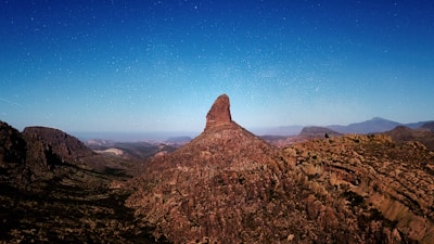 A striking landscape features a prominent rocky peak rising sharply against a backdrop of a starry night sky. Surrounding the peak, rugged terrain with numerous ridges and valleys creates a complex and dramatic natural scene.