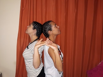 Two individuals with braided hairstyles stand back to back, interlocking their hands. One wears a striped shirt while the other sports a white shirt with a necklace. The background features an orange curtain.