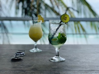 Two cocktail glasses are placed on a wooden table, with a blurred background of palm leaves and the ocean. One glass contains a yellow drink garnished with a pineapple slice, and the other has a green mint and lime drink with a black straw. Sunglasses are laid nearby on the table.