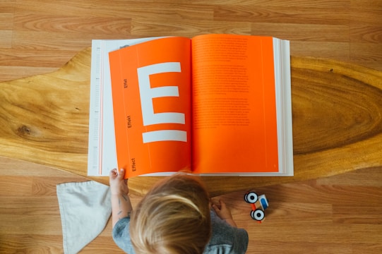 A child is viewed from above, standing on a wooden floor. In front of the child is a large open book with a bright orange page featuring white text and large white geometric shapes. Next to the child is a small wooden toy vehicle. A white cloth is also visible on the floor.