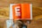 A child is viewed from above, standing on a wooden floor. In front of the child is a large open book with a bright orange page featuring white text and large white geometric shapes. Next to the child is a small wooden toy vehicle. A white cloth is also visible on the floor.
