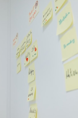 Sticky notes with handwritten words and diagrams are arranged on a white wall. The text on the notes includes terms related to design, like 'Product Design', 'Web Design', 'Branding', 'UX', and 'IA'. Different colored dots and small drawings are present alongside the text.