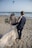A couple walks hand in hand on a sandy beach, dressed in wedding attire. The woman is wearing a lace wedding gown and holding a bouquet, while the man is in a navy blue suit. They are facing the ocean, with waves gently crashing on the shore. In the sky, there are a few kites flying, adding a colorful touch to the serene setting.