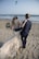 A couple walks hand in hand on a sandy beach, dressed in wedding attire. The woman is wearing a lace wedding gown and holding a bouquet, while the man is in a navy blue suit. They are facing the ocean, with waves gently crashing on the shore. In the sky, there are a few kites flying, adding a colorful touch to the serene setting.