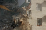 A construction excavator with a bucket attachment is demolishing a brick building. Rubble and dust are visible as the bricks fall and the structure collapses. The building appears to be residential with windows and parts of the interior exposed.