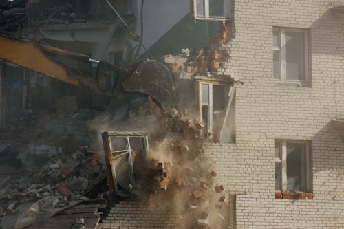 A construction excavator with a bucket attachment is demolishing a brick building. Rubble and dust are visible as the bricks fall and the structure collapses. The building appears to be residential with windows and parts of the interior exposed.