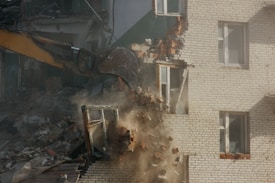 A construction excavator with a bucket attachment is demolishing a brick building. Rubble and dust are visible as the bricks fall and the structure collapses. The building appears to be residential with windows and parts of the interior exposed.
