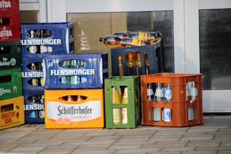Several colored crates filled with various bottles are stacked and arranged outside. The bottles appear to contain drinks, with labels indicating different brands and types. The crates are in shades of blue, orange, green, and more, with some logos and text visible on them.