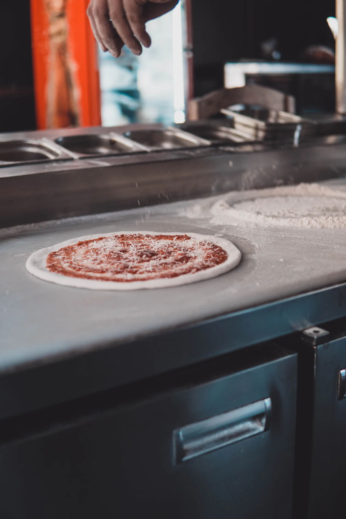 A hand-pressed pizza, sauced and cheesed to order, resting on the prep counter with the topping wells behind it