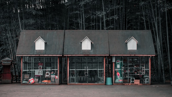 Three small wooden cabins with glass fronts are positioned side by side against a backdrop of dense bamboo forest. The cabins have steeply pitched roofs with small dormer windows. Various products and objects are visible through the glass, including a vending machine, beverage containers, and different packaged items. The scene exudes a rustic and somewhat nostalgic feel.