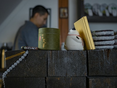 A collection of items on a dark wooden counter includes a green cylindrical container with Chinese characters, a white ceramic figure resembling a cat, a stack of round packages wrapped in paper, and yellow rectangular sheets. A string of round beads is draped over the front edge of the counter. In the background, a blurred figure of a person is visible, suggesting an indoor setting.