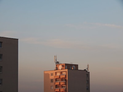 Two apartment buildings under a clear sky during sunset. The building on the right has several antennas and satellite dishes on the roof, with warm sunlight casting a golden hue on its facade. The sky is mostly clear with subtle gradients of blue and pink.