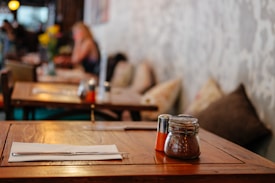 A wooden table set in a cozy dining environment with a napkin and cutlery placed neatly. Small jars for condiments are visible on the table. A blurred background reveals more tables, chairs, cushions, and a dimly lit atmosphere, suggesting a relaxed setting.