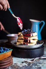 person holds spoons with berries near waffles on plate