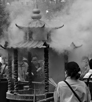 A group of people stand around a traditional incense burner, thick with smoke. The structure has ornate details and a tiered, pagoda-like roof. Individuals are holding incense sticks, engaged in a ritual or ceremony.