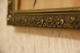 A close-up view of an ornate picture frame. The frame features intricate carvings and grooves, with a brass or golden finish. The background consists of a textured wall with a subtle floral pattern in light hues.